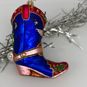 Op de afbeelding: Blauwe glazen kerstversiering in de vorm van een cowboylaars met roze, rode en gouden accenten. De laars heeft een ster en een vleugelontwerp aan de zijkant. De versiering heeft een gouden label met het woord "Polen" erop.