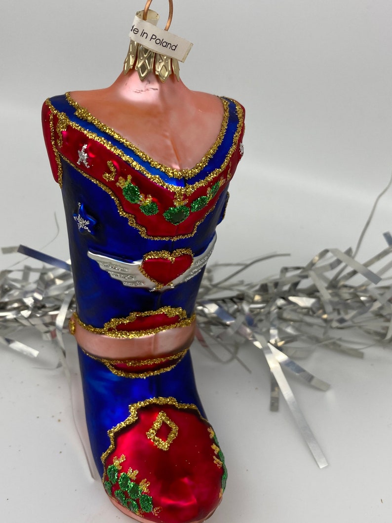 Op de afbeelding: Een blauwe en rode glazen kerstversiering in de vorm van een cowboylaars met gouden glitteraccenten en een hart met vleugels. De versiering heeft een label met de tekst "Made in Poland".