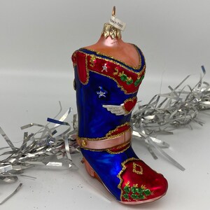 Op de afbeelding: Een blauwe en rode glazen kerstversiering in de vorm van een cowboylaars. De laars heeft gouden versieringen en een hart met vleugels aan de zijkant. De versiering heeft een label met de tekst "Made in Poland".
