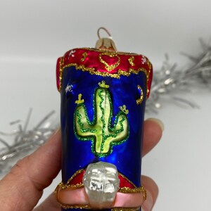 Op de afbeelding: Een roze en blauwe glazen kerstversiering in de vorm van een cowboylaars met een cactusontwerp. De laars heeft een rode en gouden rand en een zilveren sporen.