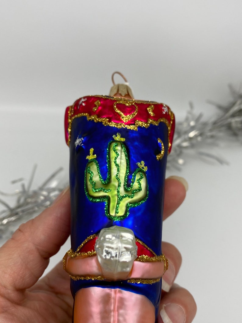 Op de afbeelding: Een roze en blauwe glazen kerstversiering in de vorm van een cowboylaars met een cactusontwerp. De laars heeft een rode en gouden rand en een zilveren sporen.