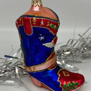 Op de afbeelding: Een blauwe en rode glazen kerstversiering in de vorm van een cowboylaars. De laars heeft gouden glitteraccenten en een witte ster aan de zijkant. De versiering heeft een gouden lus aan de bovenkant om op te hangen.