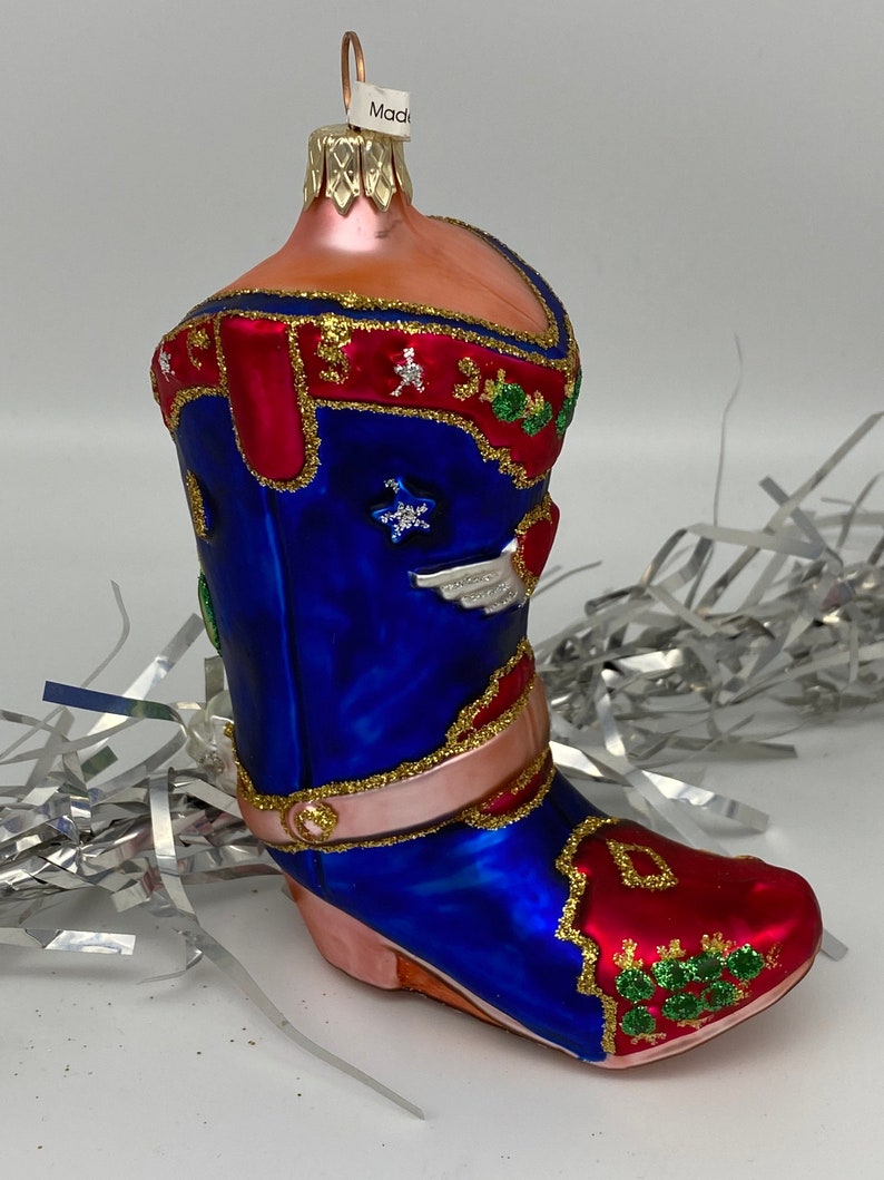 Op de afbeelding: Een blauwe en rode glazen kerstversiering in de vorm van een cowboylaars. De laars heeft gouden glitteraccenten en een witte ster aan de zijkant. De versiering heeft een gouden lus aan de bovenkant om op te hangen.