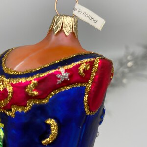 Op de afbeelding: Een blauwe en rode glazen kerstbal met gouden versiering en een wit label met de tekst "Gemaakt in Polen".