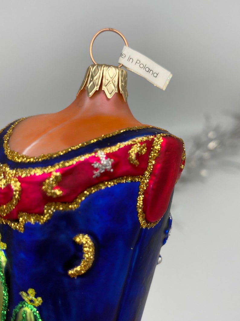 Op de afbeelding: Een blauwe en rode glazen kerstbal met gouden versiering en een wit label met de tekst "Gemaakt in Polen".