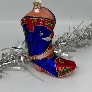 Op de afbeelding: Een blauwe en rode glazen kerstversiering in de vorm van een cowboylaars. De laars heeft gouden versieringen en groene en witte details. De versiering heeft een gouden lus aan de bovenkant om op te hangen.