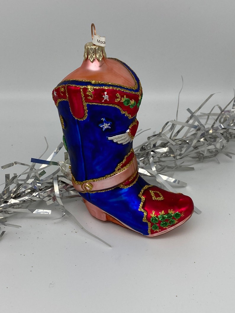 Op de afbeelding: Een blauwe en rode glazen kerstversiering in de vorm van een cowboylaars. De laars heeft gouden versieringen en groene en witte details. De versiering heeft een gouden lus aan de bovenkant om op te hangen.