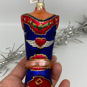 Op de afbeelding: Een blauwe en rode glazen kerstversiering in de vorm van een cowboylaars met gouden glitteraccenten. De laars heeft een hart met vleugels aan de voorkant en een rood en gouden ontwerp op de hiel. De versiering heeft een gouden koord om op te hangen.