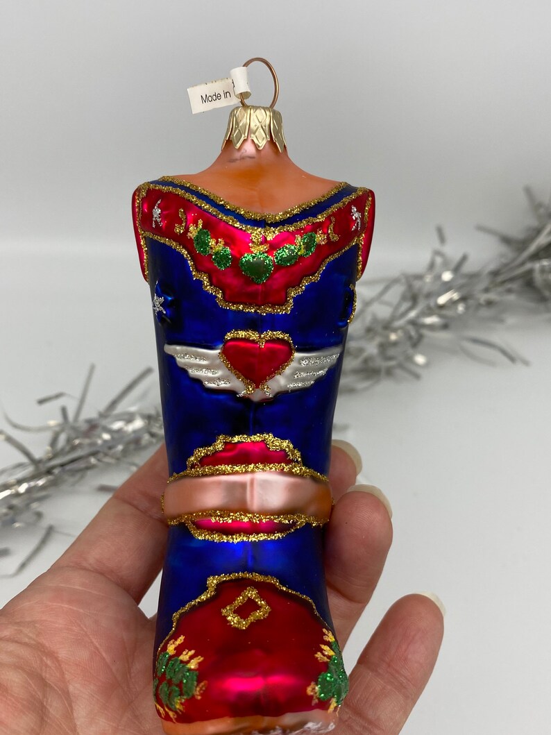 Op de afbeelding: Een blauwe en rode glazen kerstversiering in de vorm van een cowboylaars met gouden glitteraccenten. De laars heeft een hart met vleugels aan de voorkant en een rood en gouden ontwerp op de hiel. De versiering heeft een gouden koord om op te hangen.