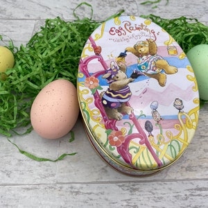 May include: A vintage oval tin box with a colorful illustration of two teddy bears painting Easter eggs. The text on the box reads "Egg Painting's "Walking in Eggy Shells".