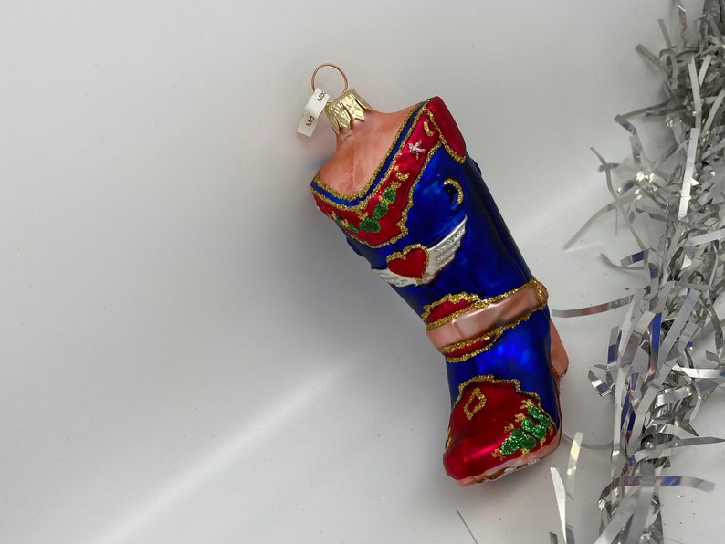 Op de afbeelding: Een glazen kerstversiering in de vorm van een cowboylaars. De laars is versierd met rode, blauwe en groene verf en heeft een hart met vleugels, een cactus en een gouden ster. De versiering heeft een gouden koord om op te hangen.
