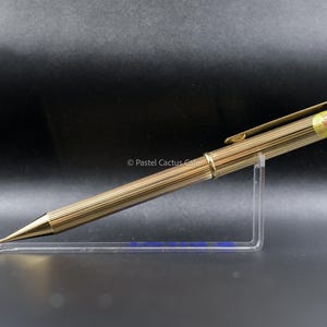 以下が含まれることがあります： リブ加工が施されたゴールドカラーのペン。クリップとテキストの小さなラベルが付いています。ペンは、暗い背景に対して透明なアクリルスタンドに展示されています。ペンのデザインはエレガントでクラシックです。