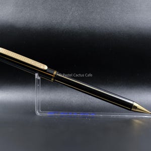 Peut inclure: Un stylo à bille noir avec des accents dorés, doté d'un clip doré texturé et d'une pointe dorée effilée. Le stylo est présenté sur un support en acrylique transparent. Le texte Pastel Cactus Cafe est visible au centre de l'image.