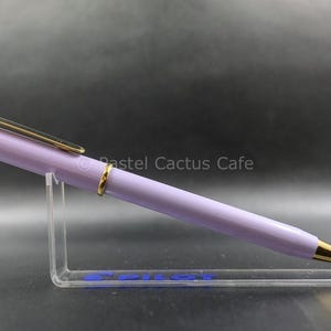 Op de afbeelding: Een lavendelkleurige pen met goudkleurige details, waaronder de punt, clip en ringen. De pen wordt gepresenteerd op een transparante acryl standaard. De pen heeft een strak, modern design.