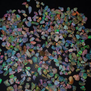 May include: A large collection of iridescent opal chips in various shades of blue, green, red, and orange. The chips are scattered on a black background.
