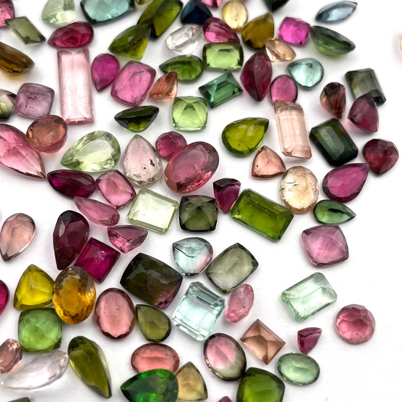 Op de afbeelding: Een verzameling losse edelstenen in verschillende vormen en maten, waaronder groen, roze en blauw. De edelstenen zijn verspreid over een wit oppervlak.