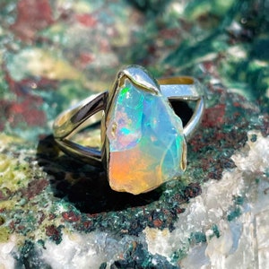 May include: A silver ring with a raw, iridescent opal gemstone. The opal has a rough, natural shape and displays a rainbow of colors.