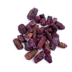 Op de afbeelding: Een verzameling ruwe robijn edelstenen, met een dieprode kleur en verschillende vormen en maten. De robijnen zijn gerangschikt op een witte achtergrond.