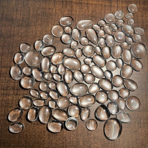 May include: A collection of clear, smooth, oval-shaped stones. The stones are arranged in a loose, organic pattern on a brown surface.