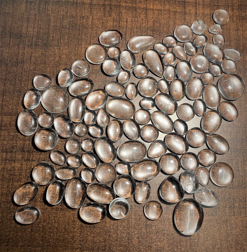 May include: A collection of clear, smooth, oval-shaped stones. The stones are arranged in a loose, organic pattern on a brown surface.