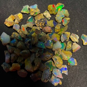 May include: A collection of rough opal stones, showcasing a variety of colors including blues, greens, reds, and yellows. The raw gemstones have a natural, earthy appearance with irregular shapes and sizes. The opals are displayed against a dark background.