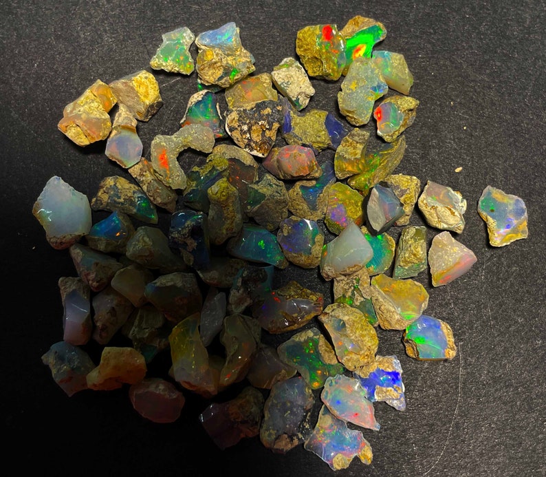 May include: A collection of rough opal stones, showcasing a variety of colors including blues, greens, reds, and yellows. The raw gemstones have a natural, earthy appearance with irregular shapes and sizes. The opals are displayed against a dark background.