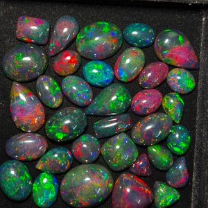 May include: A collection of iridescent black opals with green, blue, and red flashes. The opals are various shapes and sizes and are arranged in a black velvet box.