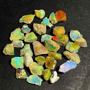 Puede incluir: Una colección de piedras preciosas de ópalo en bruto, que muestra una variedad de colores, incluyendo verde, amarillo, naranja y azul. Los ópalos tienen texturas naturales rugosas y están dispersos sobre un fondo oscuro. Algunas piedras exhiben un vibrante juego de colores.