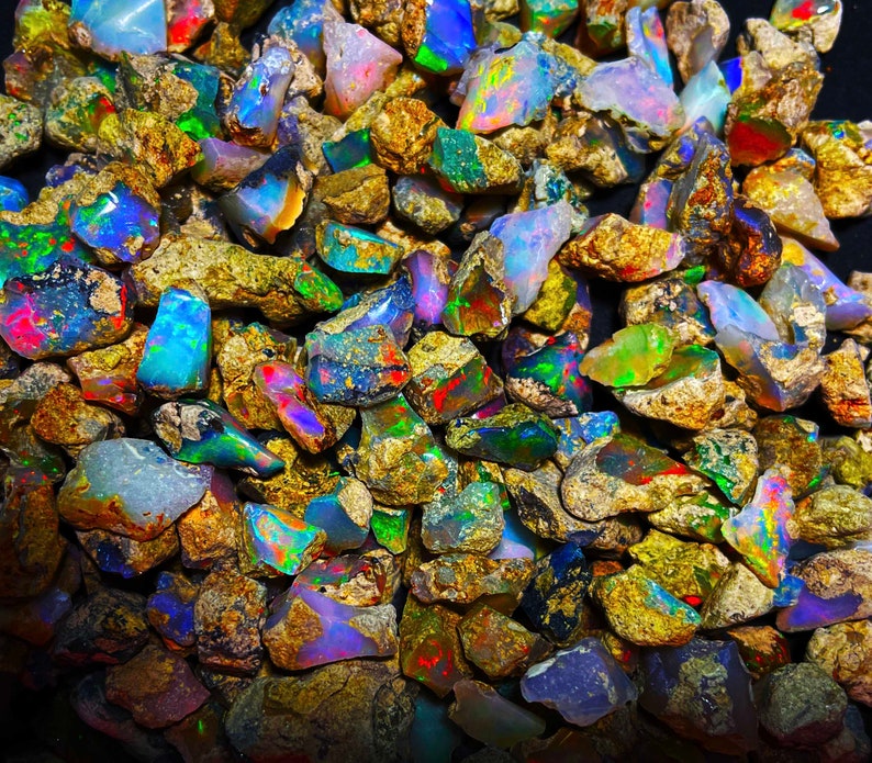 May include: A close-up view of a pile of rough opal gemstones. The opals display a variety of colors, including blue, green, red, and yellow, with a shimmering, iridescent effect. The opals are surrounded by a matrix of brown rock.