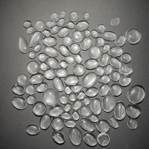 May include: A collection of clear, smooth, oval-shaped glass stones. The stones are various sizes and are scattered on a gray surface.
