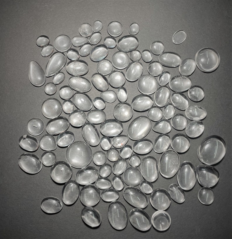 May include: A collection of clear, smooth, oval-shaped glass stones. The stones are various sizes and are scattered on a gray surface.