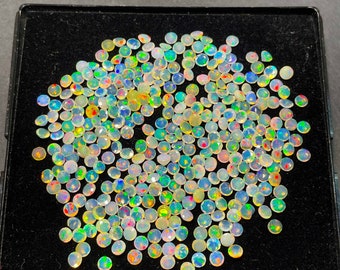 May include: A collection of small, round, faceted opals in a black tray. The opals display a variety of colors, including white, yellow, green, blue, and red, with a shimmering, iridescent effect. The gemstones are closely packed together.