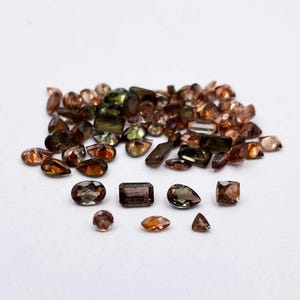 May include: A collection of loose brown, green, and black gemstones. The gemstones are various shapes and sizes, including oval, pear, and rectangular.
