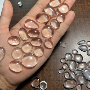 May include: A collection of clear quartz cabochons of various sizes and shapes, displayed on a hand. The cabochons are smooth and polished, with a translucent appearance.