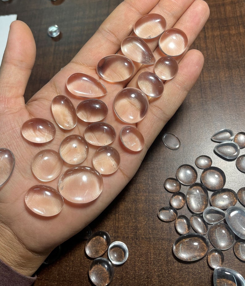 May include: A collection of clear quartz cabochons of various sizes and shapes, displayed on a hand. The cabochons are smooth and polished, with a translucent appearance.