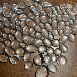 May include: A large group of clear, smooth, oval-shaped stones. The stones are scattered on a brown surface.