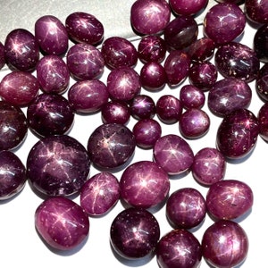 May include: A collection of loose star ruby gemstones. The rubies are a deep red colour with a star-shaped pattern in the centre of each stone. The stones are various sizes and shapes.