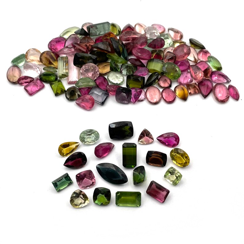 Op de afbeelding: Een grote verzameling groene, roze en bruine edelstenen. De edelstenen zijn in verschillende vormen en maten, waaronder ovaal, rond en rechthoekig. De edelstenen zijn op een witte achtergrond in een stapel gerangschikt.