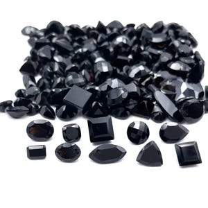 Pode incluir: Uma coleção de pedras preciosas pretas em várias formas, incluindo ovais, quadradas e em forma de gota. As pedras estão espalhadas sobre uma superfície branca, com um monte de pedras menores e irregulares no fundo.