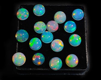 May include: A collection of round, iridescent opal gemstones in various shades of blue, green, and pink. The gemstones are displayed in a black velvet-lined box.