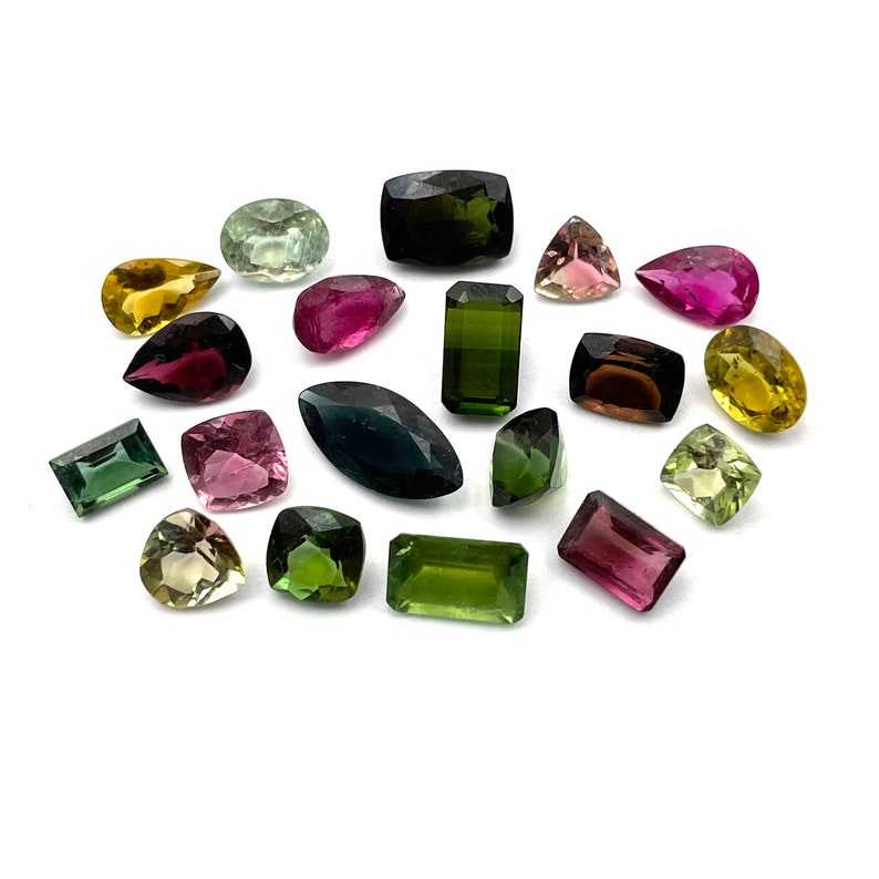 Op de afbeelding: Assortiment van groene, roze en gele edelstenen in verschillende vormen en maten. De edelstenen zijn gerangschikt op een witte achtergrond.