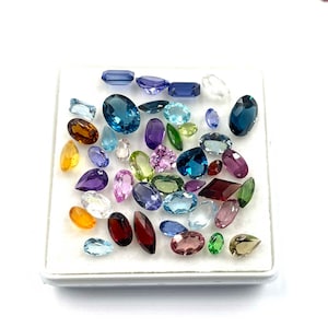 May include: A collection of various gemstones in different shapes and colors, including blue, purple, green, yellow, and red. The gemstones are displayed on a white surface, showcasing their facets and clarity. The assortment includes oval, pear, and rectangular cuts.