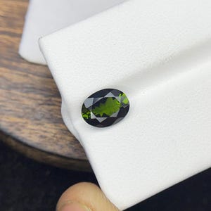 May include: An oval-cut, dark green gemstone, likely a tourmaline or similar gem, displayed on a white surface. The gem is held by a person's fingers, showcasing its polished facets and rich color. The gemstone is approximately 0.5 inches in length.