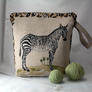 May include: A beige zippered knitting bag with a zebra design and a brown handle. Two balls of green yarn are in the foreground.