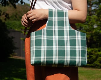 May include: A green and white plaid tote bag with a rectangular shape and a white interior. The bag is held by a person wearing a white t-shirt and orange pants. Two wooden knitting needles are held in the other hand.