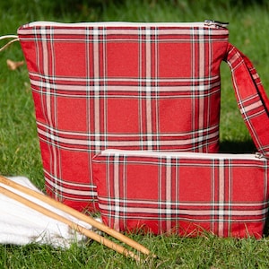May include: Two red and white plaid zippered pouches with a white yarn ball and wooden knitting needles on a green grassy surface.