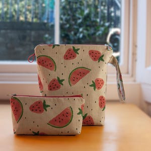 May include: Two beige zippered pouches with a watermelon and strawberry print. The larger pouch has a handle. Both pouches have red zipper pulls.