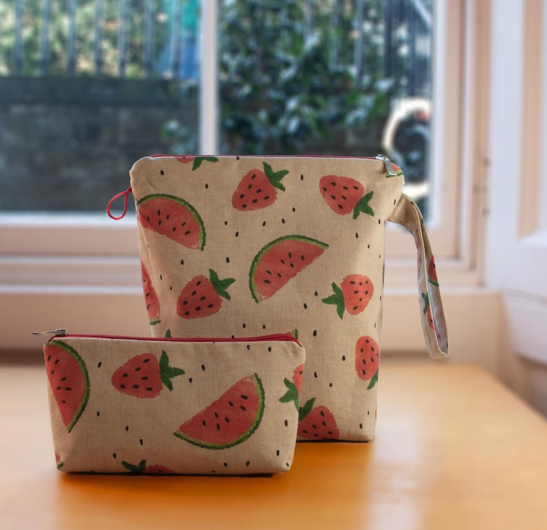 May include: Two beige zippered pouches with a watermelon and strawberry print. The larger pouch has a handle. Both pouches have red zipper pulls.