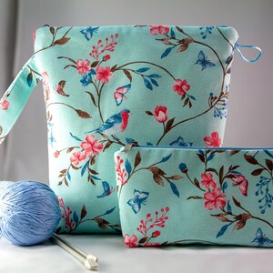 May include: Two teal blue zippered pouches with a floral print featuring pink flowers, brown branches, and blue birds. The larger pouch has a handle and the smaller pouch is lying on a white surface.