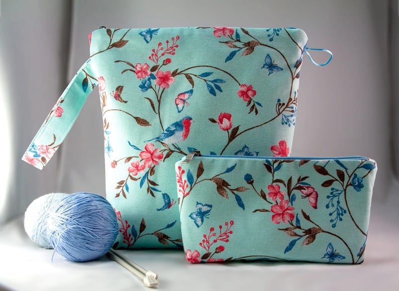 May include: Two teal blue zippered pouches with a floral print featuring pink flowers, brown branches, and blue birds. The larger pouch has a handle and the smaller pouch is lying on a white surface.